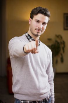 Handsome young man pointing finger at you, smiling. Indoors shot inside a house