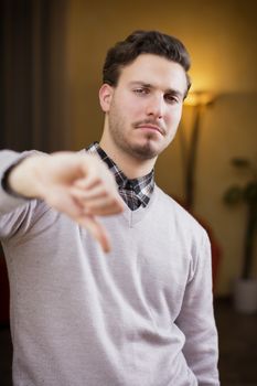 Disappointed or displeased young man doing thumb down sign. Indoors shot inside a house