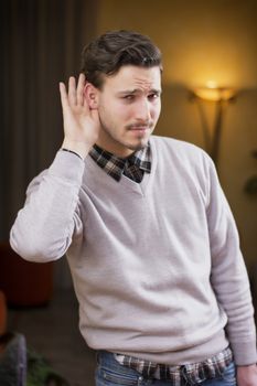 Handsome young man can't hear, putting hand around his ear. Indoors shot inside a house