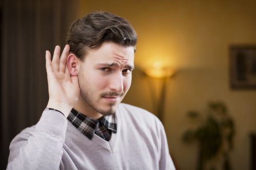 Handsome young man can't hear, putting hand around his ear. Indoors shot inside a house