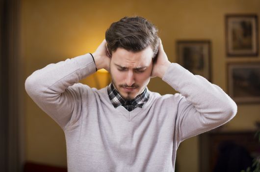 Handsome young man covering his ears, stressed or unhappy because of too much noise. Indoors shot