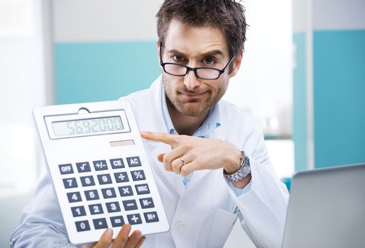 Young friendly doctor pointing at a big calculator.