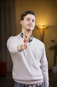 Attractive young man doing peace or victory sign with two fingers, indoor shot inside a house