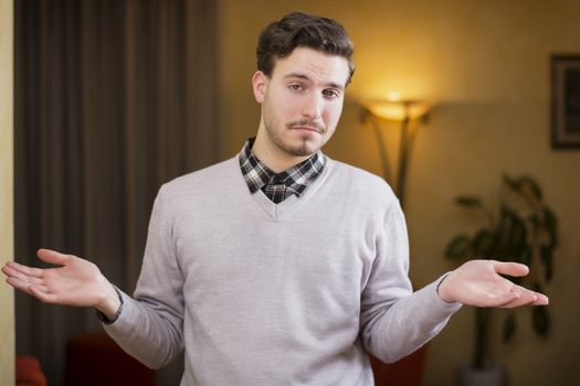 Confused or doubtful young man shrugging with palms open, indoor shot in a house