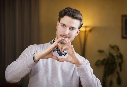 Handsome young man making heart sign with his hands and fingers. Indoors shot inside a house