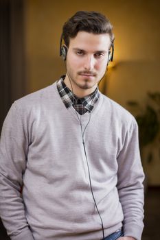 Attractive young man listening to music on headphones at home, looking at camera, serious
