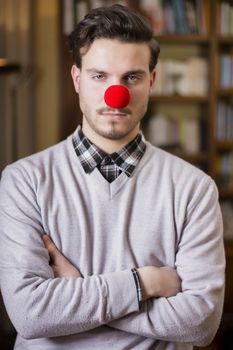 Serious young man with red clown nose, arms crossed, indoor shot