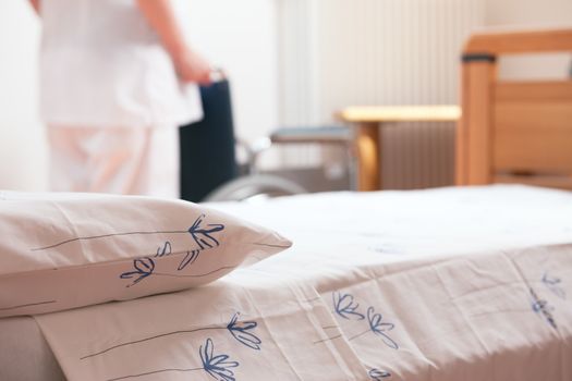 Bed at nursing home with nurse and wheelchair on background.