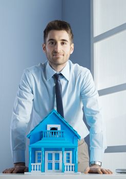 Young real estate agent leaning on desk with model house.