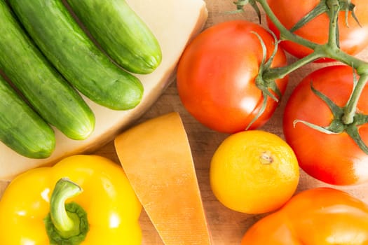 Colourful background of healthy fresh groceries with a close up overhead view of baby green cucumbers, yellow sweet pepper, ripe red grape tomatoes, a lemon and wedge of cheese with an orange rind