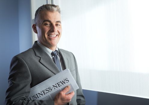 Confident businessman holding a newspaper and smiling next to office window.