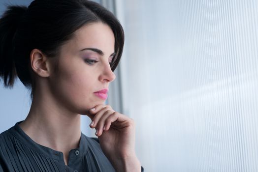 Young pensive woman at window looking away with hand on chin.