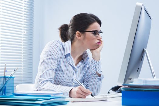 Bored office worker at desk staring at computer screen with hand on chin.