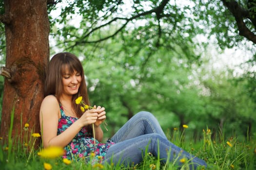 Teenage girl is resting in the park, sitting on the grass. Enjoying the nature and playing with dandelions