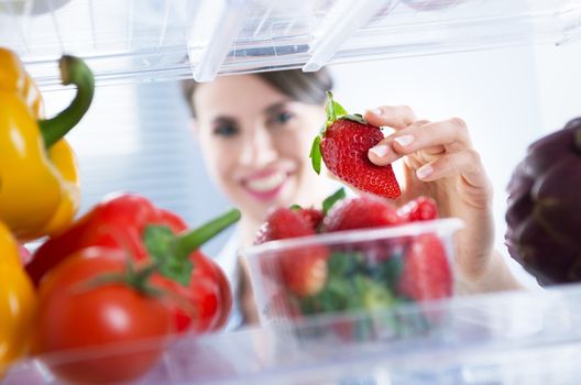 Young woman smiling and taking a fresh strawberry from refrigerator.