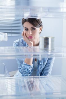 Young sad woman looking at one tin in her empty fridge.