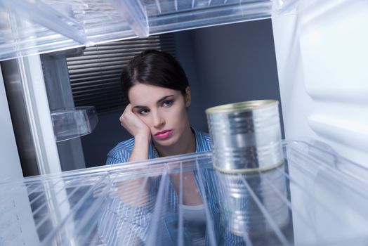 Young sad woman looking at one tin in her empty fridge.