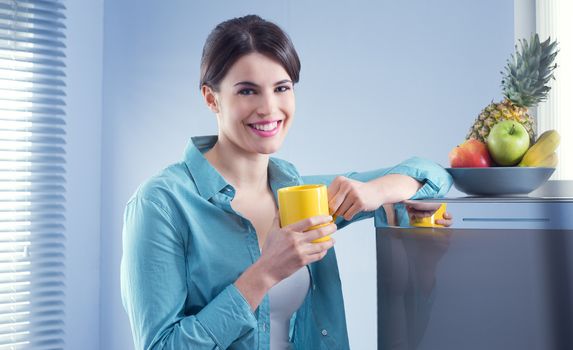 Young cheerful woman holding a yellow mug and leaning against refrigerator.