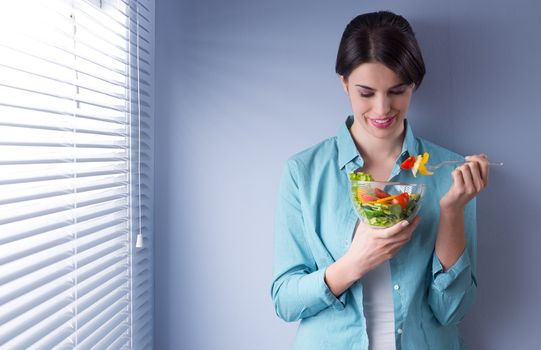 Woman smiling and eating salad in front of a window.