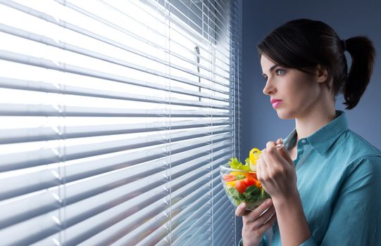 Pensive woman eating healthy salad in front of a window.