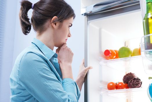 Young pensive woman looking in the refrigerator with hand on chin.