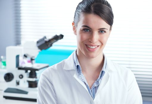 Confident female researcher in the chemistry lab with microscope and laboratory glassware on background.