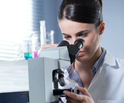 Young female researcher using microscope in the chemistry lab with laboratory glassware on background.