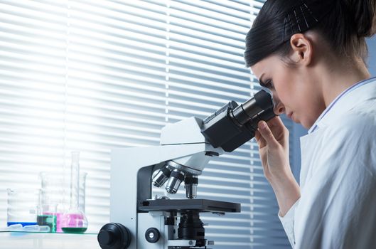 Young female researcher using microscope in the chemistry lab with laboratory glassware on background.