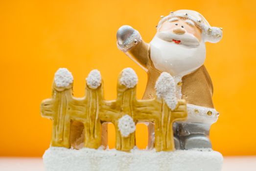 Yellow old time Santa Claus figurine standing behind garden fence and waving with one hand, shot in studio, orange background