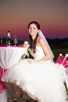Bride is sitting at the wedding table outdoors with champagne and flower in the evening glow