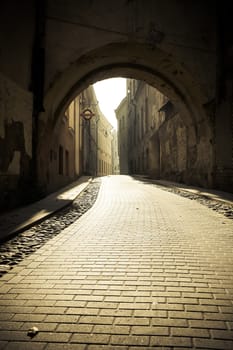 Fragment of the cobbled old town street, in the sunlight in the down, empty street, visible arch of the gate above the street.
