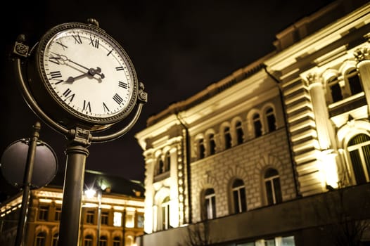 The photo presents a fragment of the night, illuminated street with tenement building in th background, and lit old tyle street clock in the front.