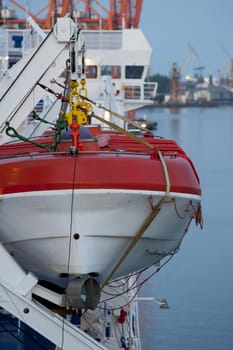 White and red lifeboat, visible ropes and elements of the lift, in the background port, cranes and sea.