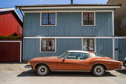 Photo presents on old fashioned car in the red colour, with red chrome rims and bumpers, small blue wooden  house with white window frames in the background, scandinavian style.