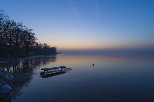 Frozen lake in winter, after sunset, with unfinished path/bridge and coastline with trees.