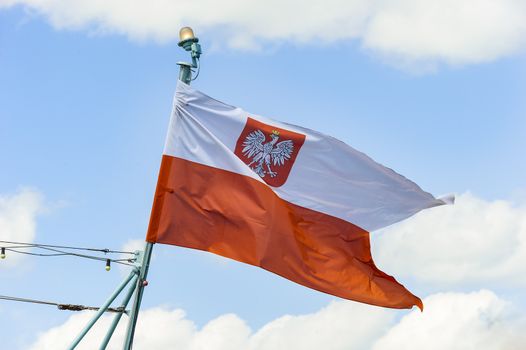 A traditional Polish red and white  flag with a crowned eagle, waving in the blue sky with white clouds.