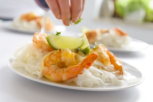 Preparation of shrimp and rice pasta meal on a white plate, visible fingers of a young woman and lime, shallow DOF.