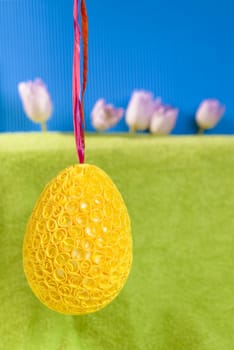 Photo presents big Easter decorative egg hanging on purple ribbon, background imitating green grass and blue sky, in grass small pink tulips visible, provided space for text.