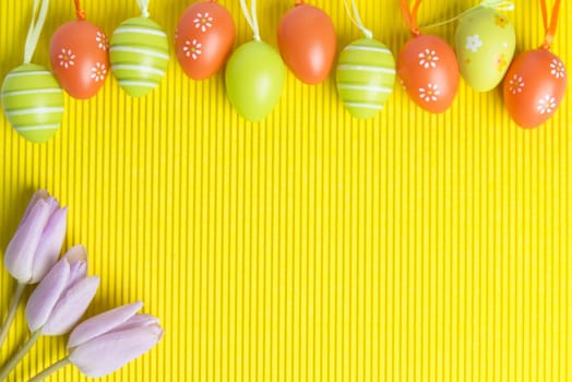 Easter arrangement on yellow background, visible orange and green  decorated eggs and pink tulips in left corner, space for text provided.