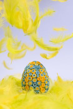 Funny, amusing and joyful photo of yellow and blue Easter egg lying in yellow feathers, visible feather falling down.
