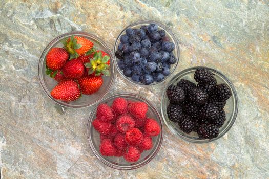 Overhead view of a selection of different fresh berries in glass jars including succulent ripe strawberries, blackberries, blueberries and raspberries for a healthy snack or dessert on a stone counter