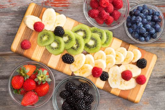 Preparing a healthy fruit salad with mixed berries in individual containers, sliced banana and exotic kiwifruit on a striped wooden board , view from above