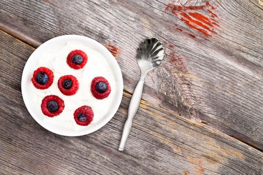 Bowl of fresh raspberries and blueberries with creamy yogurt for a healthy breakfast or daytime snack, view from above on a rustic weathered textured old wooden table with copyspace
