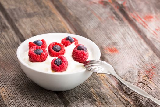 Delicious fresh raspberries and blueberries topping a bowl of creamy yogurt for a healthy snack or dessert served with a spoon on a rustic wooden picnic table, with copyspace