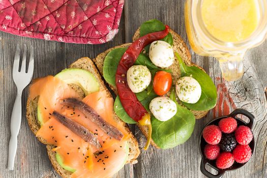 Delicious gourmet open sandwiches on wholewheat bread topped with smoked salmon, anchovies, avocado, mozzarella, baby spinach and chili pepper served on a picnic table with fresh berries