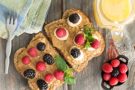 Overhead view of healthy peanut butter sandwiches on wholewheat bread topped with fresh raspberries and blackberries, herbs and mozzarella cheese on a wooden picnic table with a jug of orange juice