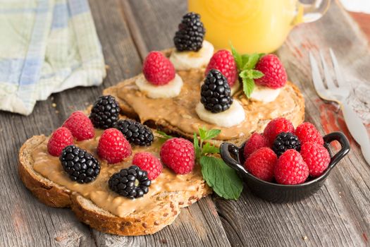 Peanut butter sandwiches on healthy wholewheat bread topped with fresh raspberries, blackberries and leafy green herbs served with a bowl of mixed berries on a picnic table