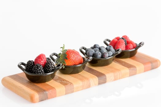 Appetizers of tasty fresh autumn berries in separate ceramic dishes with blueberries, strawberries, raspberries, and blackberries on a striped wooden board isolated on white with copyspace