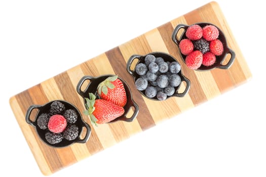 Varieties of fresh berries with raspberries, blueberries, blackberries and strawberries in individual dishes standing on a striped wooden board lying diagonally on a white background, overhead view