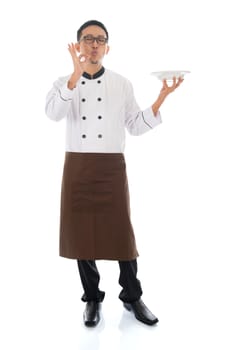 Full body Asian chef holding an empty plate, showing tasty and satisfied hand sign, standing isolated on white background.